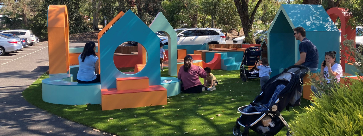 Families playing and relaxing upon colourful structures constructed in a new green area in a carpark in Croydon South