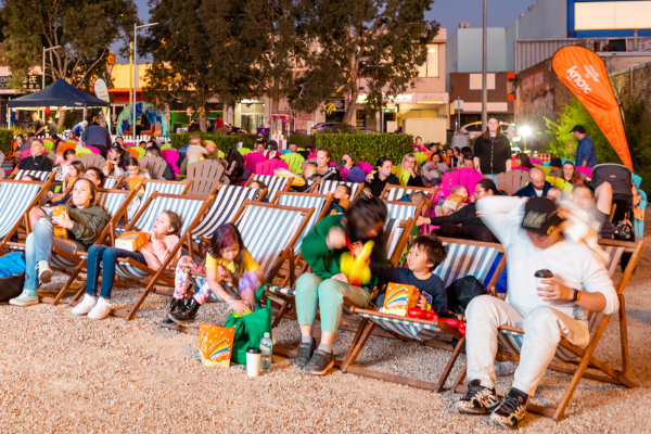 A large crowd of people are seated on outdoor chairs, facing an outdoor cinema screen (behind photographer taking this picture)