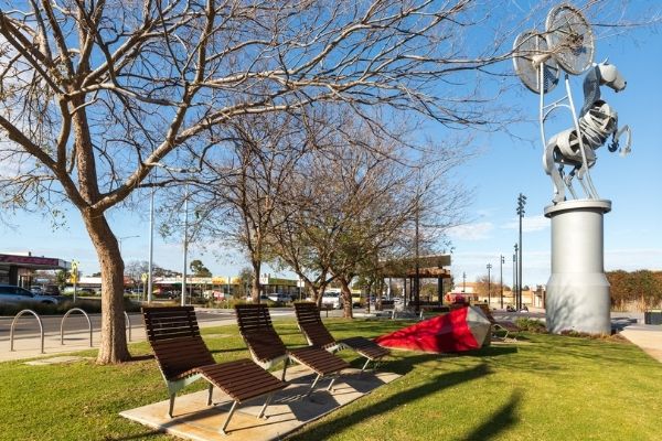 A A sunny day in Melton - this picture features public sunbeds in a park under large trees. A tall sculpture of a horse stands nearby.